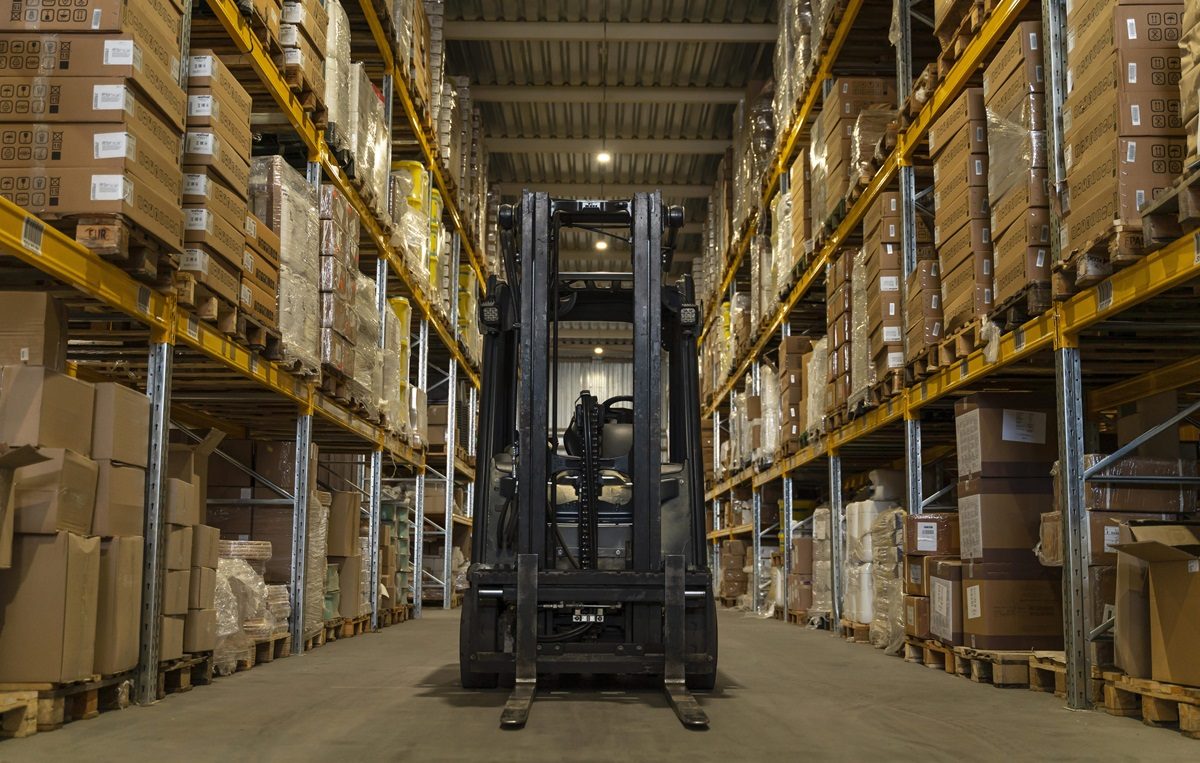 Armazém com paleteira posicionada no meio do corredor e estantes cobertas de pallets nas laterais.